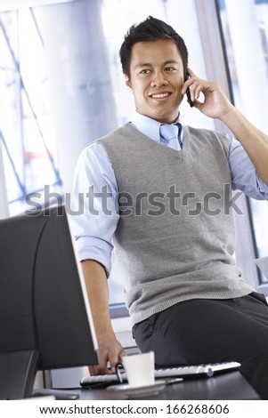 Young Asian businessman talking on mobilephone, smiling, looking away, sitting on top of desk.