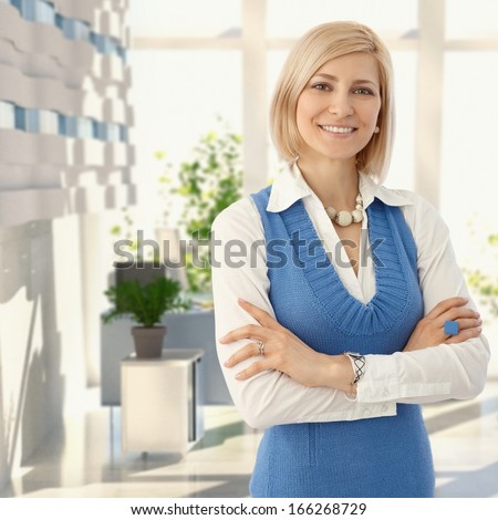 Elegant blond woman in blue standing at office smiling to camera.