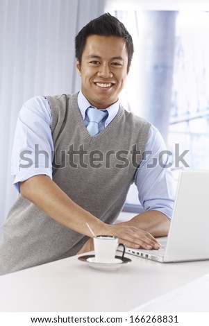 Happy young Asian businessman using laptop computer, smiling, looking at camera, leaning on counter.