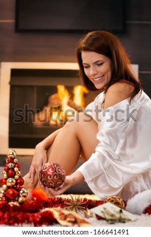 Happy attractive woman sitting by cosy fireplace, arranging christmas decoration, smiling.