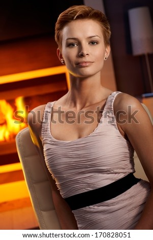 Beautiful young woman sitting by fireplace in cosy room, smiling.