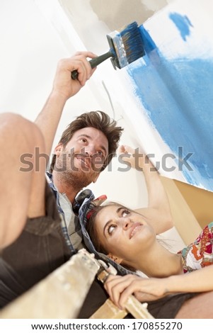Happy young couple painting wall, renovating home, smiling. Man on top of ladder. Below view.