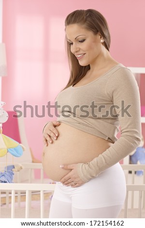 Happy pregnant woman in pink baby\'s room showing naked belly.