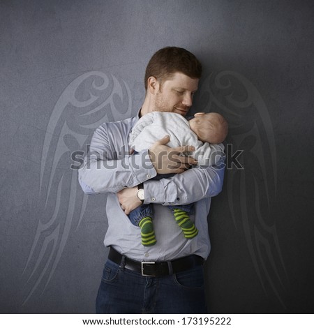 Father leaning against wall with painted angel wings, holding sleeping newborn baby, smiling affectionately.