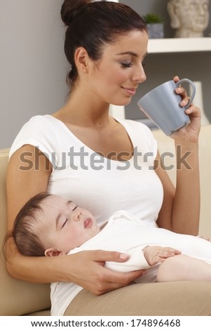 Young mother drinking tea in peace while baby sleeping in her arms.