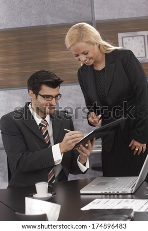 Business portrait of boss signing papers handed by secretary, both smiling.