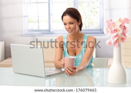 Pretty young woman using laptop computer at home, sitting at table, drinking tea.