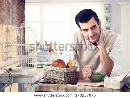 Handsome man having breakfast in trendy cottage style interior, drinking tea, reading newspaper, smiling.