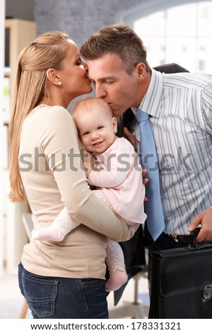 Businessman father arriving home from work, wife and little baby daughter greeting him with kiss.