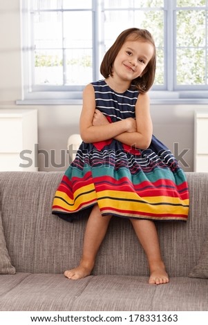 Cute little girl sitting on backrest of sofa, smiling, arms crossed.