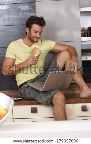 Young man sitting on kitchen counter using laptop computer, having sandwich.