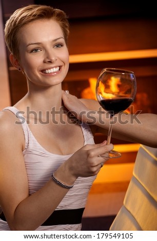 Young woman holding glass of wine smiling happy by fireplace.