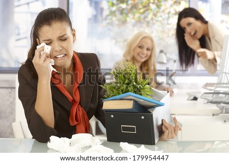 Fired young female office worker sitting at desk, crying, colleagues laughing at her at the background.