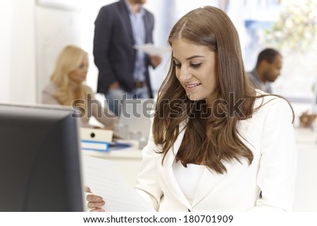 Young female office worker working in busy office, reading papers.