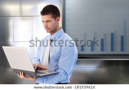 Young businessman working on laptop computer, standing by big LCD display with business diagram.