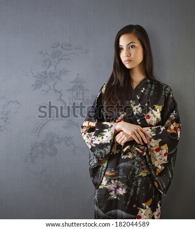 Beautiful young Japanese woman standing by grey wall, looking away, wearing kimono.