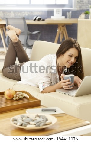Young woman drinking tea, using laptop, lying on sofa at home.
