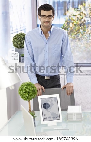 Young man standing by desk with tablet and laptop computer on it.