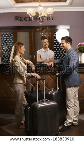 Happy young couple standing at hotel reception, checking in upon arrival, smiling.