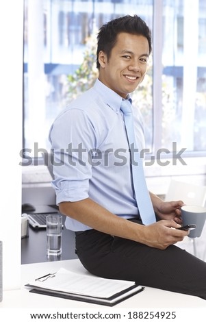 Young Asian businessman smiling as sitting on top of desk, holding mobilephone and tea mug.