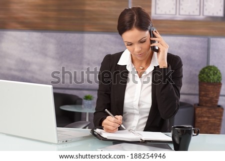 Businesswoman sitting at desk in office, talking on mobilephone, writing notes to organizer, having laptop.