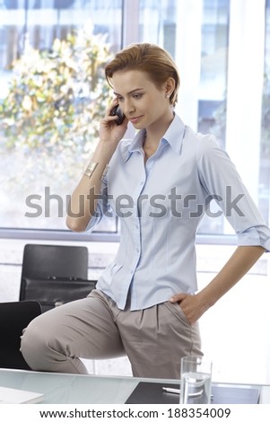 Attractive businesswoman talking on mobilephone standing at desk in office, pulling one leg up.
