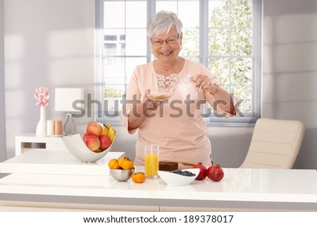 Happy old woman preparing healthy breakfast, pouring milk over cereals, smiling, looking at camera.