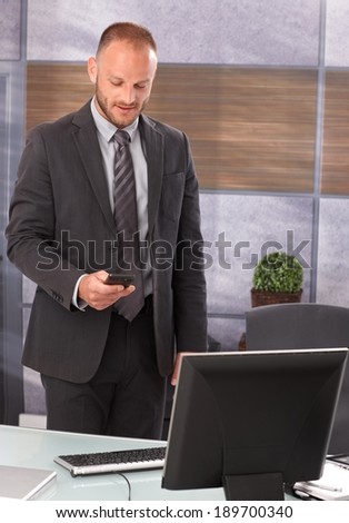 Businessman standing at desk in office, using mobilephone, dialing or writing text message.