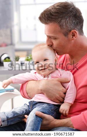 Father sitting holding on lap baby daughter, kissing smiling baby\'s head.