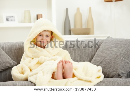 Happy smiling caucasian kid sitting in oversized bathrobe at home sofa, bare feet. Indoor, relaxed. Hood over head.