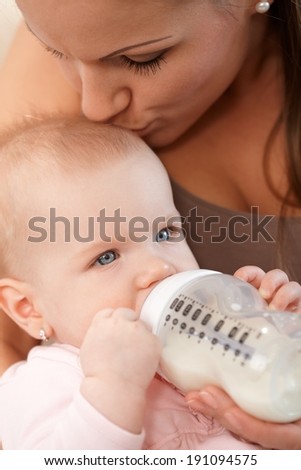 Closeup photo of mother feeding baby girl from nursing bottle, kissing head.