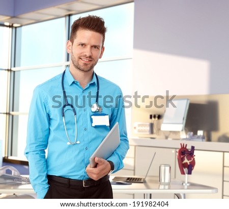 Friendly doctor standing at desk in modern office holding tablet, smiling.