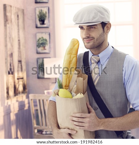 Handsome casual caucasian man holding grocery bag at vintage Paris home. Smiling, cap and french baguette bread, bristly.