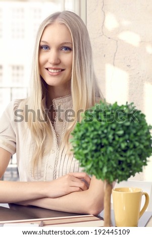 Portrait of attractive casual caucasian blonde girl sitting at home. Blue eyes, smiling, looking at camera, arms crossed.