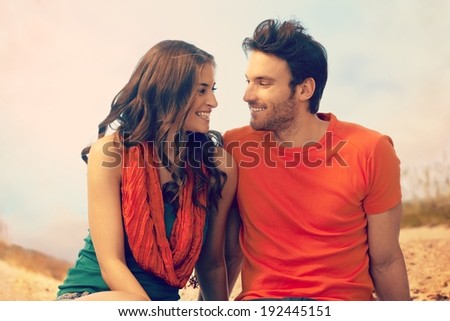 Portrait of happy romantic young casual caucasian couple sitting at outdoor holiday beach. Looking at each other, smiling. Copyspace.
