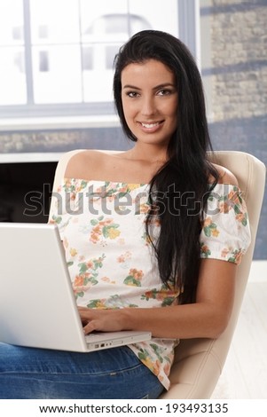 Beautiful young woman browsing internet on laptop computer, smiling, looking at camera.