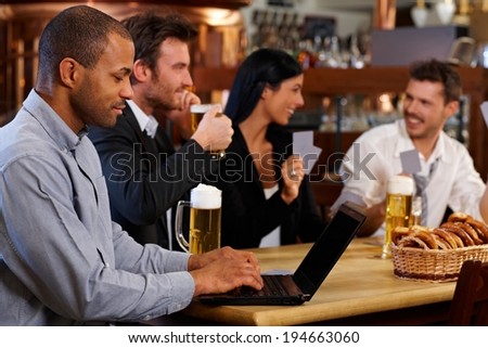 Young man browsing internet using laptop in pub, looking at screen. Friends drinking beer at background.