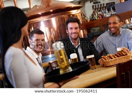 Happy young men sitting at table in pub in pub looking at waitress holding beer, smiling.