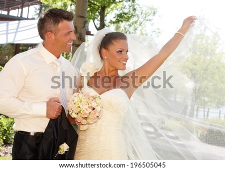 Happy couple on wedding-day, bride posing in wedding gown holding long veil.