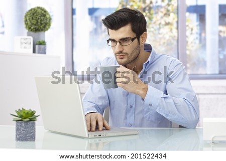 Young man sitting at desk at home, working on laptop computer, drinking coffee.
