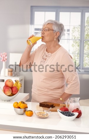 Elderly woman drinking orange juice, preparing healthy breakfast with brown bread and fruits.