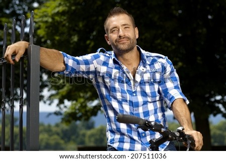 Portrait of happy, stubbly, caucasian, casual man on bicycle outdoor, leaning against fence. Smiling.