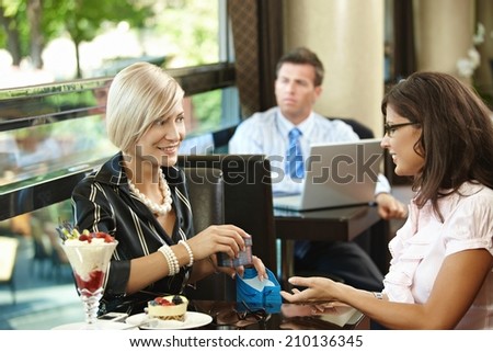 Elegant blonde caucasian businesswoman giving gift at restaurant. Sitting at table, smiling, gift box in hand, ice cream on table, indoor.