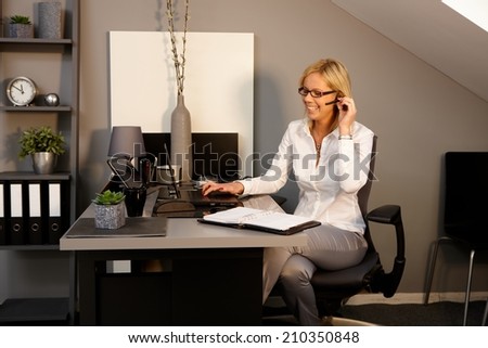 Happy blonde woman working in office, sitting at desk, using laptop computer, talking on phone.