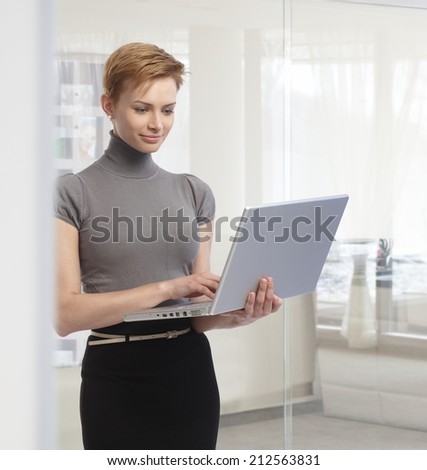 Young attractive elegant caucasian businesswoman standing with laptop computer at office. Working, looking at screen, smiling.