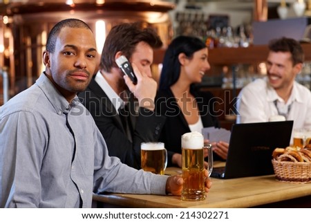 Young man drinking beer at pub with colleagues, holding mug, smiling.