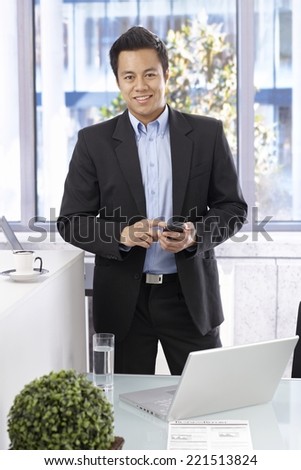 Happy smiling businessman holding mobilephone, looking at camera, standing at desk.