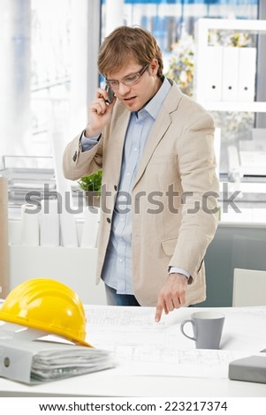 Young caucasian engineer talking on mobile phone and pointing at plan at office. Folder and hardhat on table.