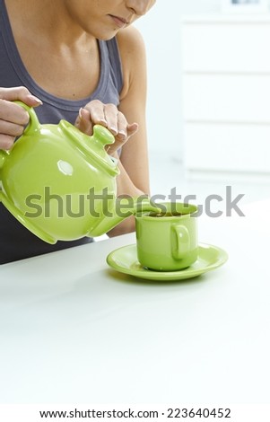 Woman pouring tea from kettle to mug on table. High key bright background, table, pastel colors, close up, indoors home. Copyspace.