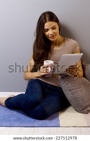 Casual young woman sitting on floor at home reading tablet, drinking tea.